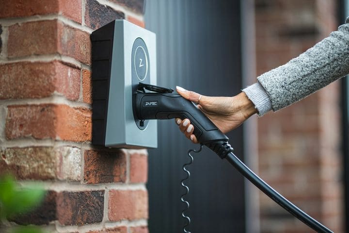 Hand lifting the Type 2 plug from a grey Zaptec charger mounted on a red brick wall