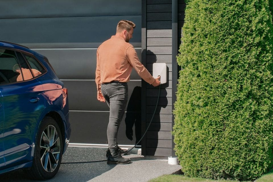 Man using a white wall charger on a dark timber-clad house, his blue electric car parked alongside on a sunny day