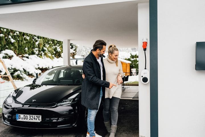 Couple standing in a carport beside their black Tesla, with a wall charger mounted on the post