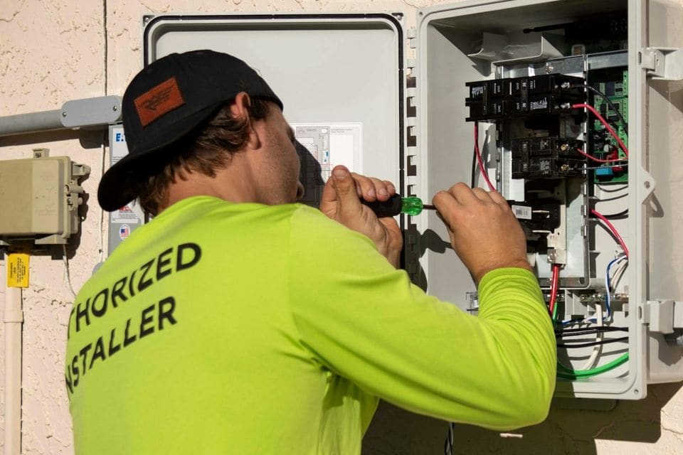 Electrician with a screwdriver wiring an open outdoor electrical panel on a house wall