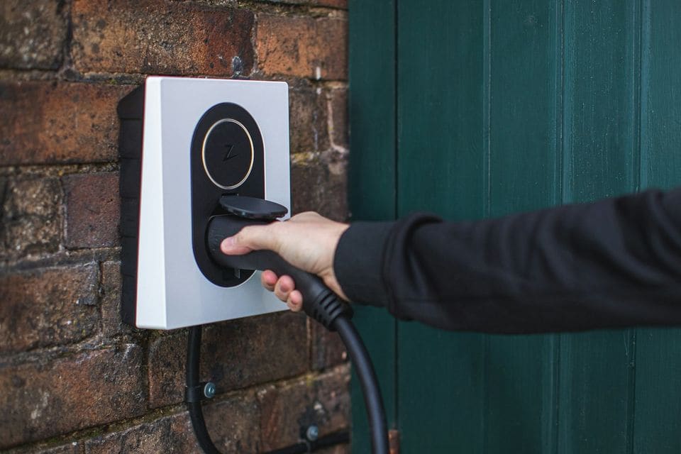Person plugging a charging cable into a white Zaptec wall charger on an old brick wall