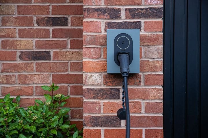 Grey Zaptec wall charger with the cable plugged in, mounted on a red brick wall beside a shrub