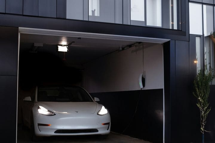 White Tesla Model 3 parked in a lit home garage at dusk, a wall charger glowing green on the side wall